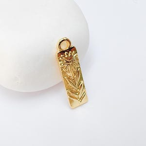May include: A gold-colored rectangular charm with a floral design. The charm features a detailed embossed pattern of a flower and leaves. It has a small loop at the top for attaching to a necklace or bracelet. The charm is displayed on a white background.