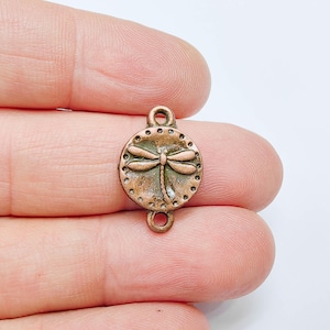 May include: A copper-colored round charm with a dragonfly design. The charm has two loops for attaching to jewelry. The dragonfly is in the center of the charm, and the border has small, raised dots.