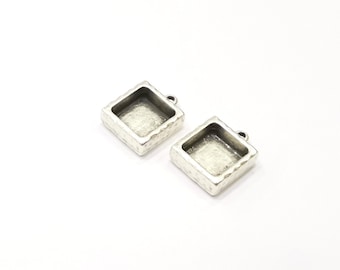 May include: Two silver-toned square pendant trays with textured edges. Each tray has a small loop for attaching to a chain or other jewelry component. The trays are empty, ready for embellishment.
