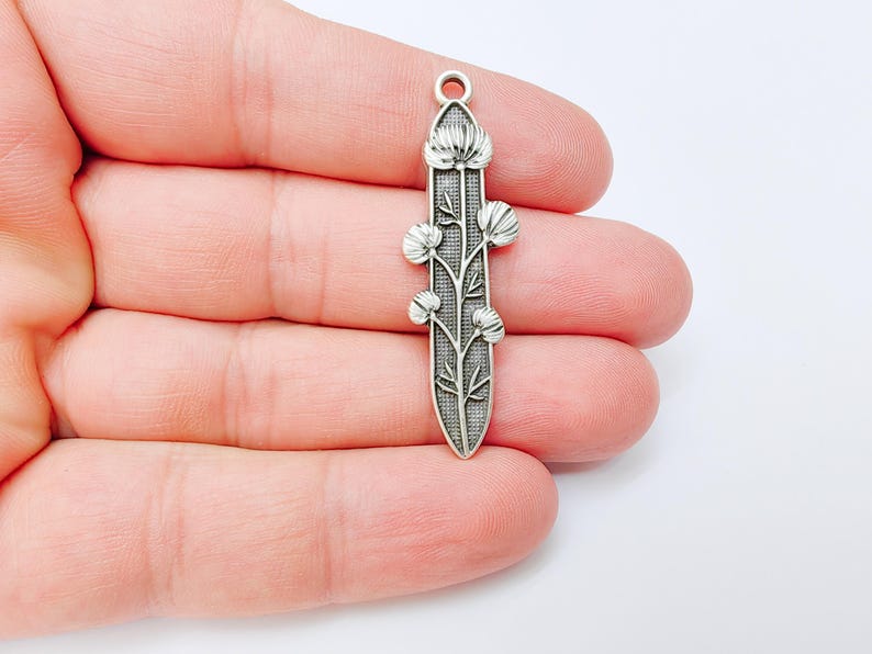 Op de afbeelding: Zilverkleurige metalen hanger met een bloemenontwerp. De hanger heeft een verticale, langwerpige vorm met een gedetailleerd patroon van bloemen en bladeren. Het ontwerp is symmetrisch en heeft een lus aan de bovenkant om op te hangen.