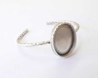 May include: A silver-toned cuff bracelet with an oval bezel setting. The bracelet has a hammered texture and an open design. The bezel is designed to hold a cabochon or other decorative element. The bracelet is shown on a white background.