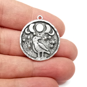 May include: Silver charm featuring a raven with three crescent moons and a full moon above it. The charm is textured and has a rustic look.