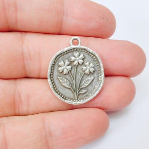 May include: A silver-toned round pendant featuring a floral design. The pendant has a raised border with small dot detailing. The central design shows three flowers with detailed leaves and stems. The pendant has a small loop at the top for attaching to a chain or cord.