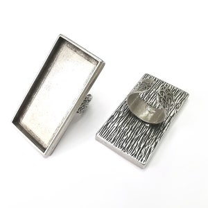 May include: Silver ring blank with a rectangular shaped bezel. The ring is made of metal and has a textured finish.