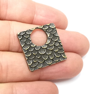 Pode incluir: Um amuleto de metal quadrado com um furo no centro. O amuleto tem uma superfície texturizada que se assemelha a escamas. O amuleto é de cor bronze.