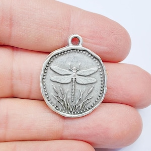 Puede incluir: Un colgante redondo plateado con un diseño de libélula. La libélula está en el centro, rodeada por un borde de cuentas, con hojas de hierba debajo. Tiene un lazo en la parte superior para sujetarlo a una cadena. Este charm es adecuado para la creación de joyas.