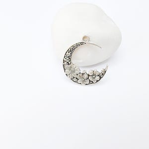 May include: A silver-toned crescent moon pendant with floral detailing. The pendant features a series of embossed flowers along the bottom curve and a small loop at the top for a chain. The pendant is displayed on a white stone.