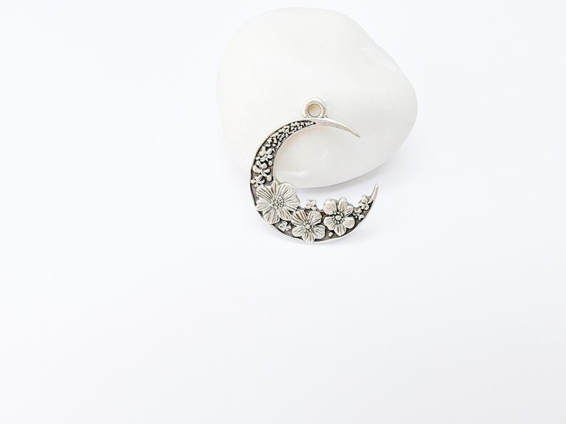 May include: A silver-toned crescent moon pendant with floral detailing. The pendant features a series of embossed flowers along the bottom curve and a small loop at the top for a chain. The pendant is displayed on a white stone.