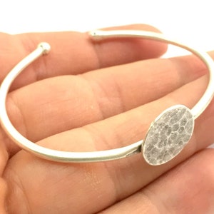 Puede incluir: Un brazalete de plata con un dije circular martillado. El brazalete está abierto y tiene un diseño simple y minimalista.
