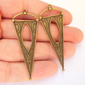 Può includere: Due ciondoli triangolari in metallo color oro antico con disegni intricati e una forma ovale ritagliata al centro.