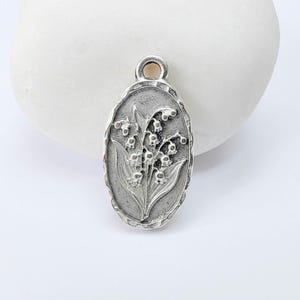 Op de afbeelding: Ovale zilveren hanger met een gedetailleerd reliëf van lelietjes-van-dalen bloemen en bladeren. De hanger heeft een getextureerde rand en een kleine lus aan de bovenkant om aan een ketting te bevestigen. Een klassiek bloemenmotief.