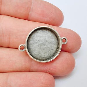 Puede incluir: Un colgante de bisel redondo plateado con un centro empotrado. El colgante tiene dos pequeños bucles a cada lado para sujetarlo a una cadena u otros componentes de joyería. El bisel se sostiene en una mano.