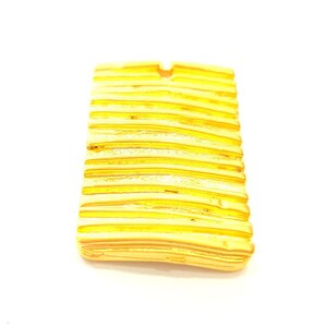 Könnte beinhalten: Ein goldfarbener Anhänger mit einem strukturierten, gestreiften Design. Der Anhänger ist rechteckig und hat eine leicht abgerundete Kante.