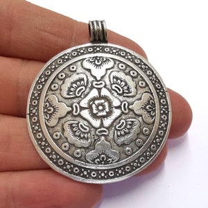 May include: A silver-colored pendant with an intricate floral design. The round pendant features a central flower surrounded by stylized petals and a border of small circles. The pendant has a loop at the top for a chain.