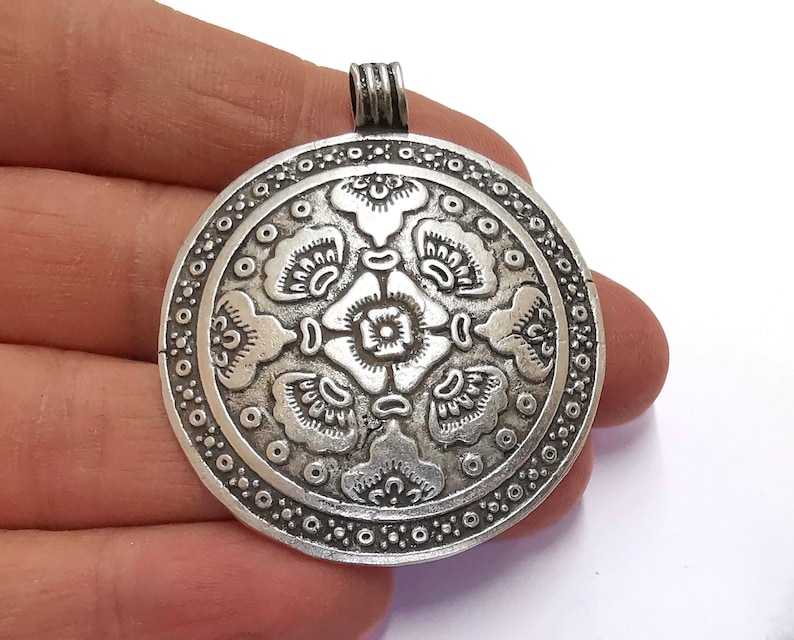 May include: A silver-colored pendant with an intricate floral design. The round pendant features a central flower surrounded by stylized petals and a border of small circles. The pendant has a loop at the top for a chain.