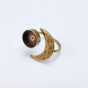 Op de afbeelding: Een messingkleurige ring met een halve maan en een cirkelvormige zetting. De maan heeft een getextureerd oppervlak en de band van de ring buigt om de vinger. De zetting is ontworpen om een klein object vast te houden.