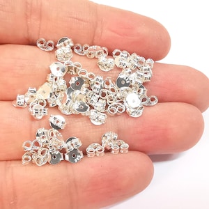 May include: A pile of silver earring backs. The backs are made of metal and have a circular shape with a small hole in the center. They are designed to be used with stud earrings.