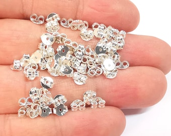 May include: A pile of silver earring backs. The backs are made of metal and have a circular shape with a small hole in the center. They are designed to be used with stud earrings.