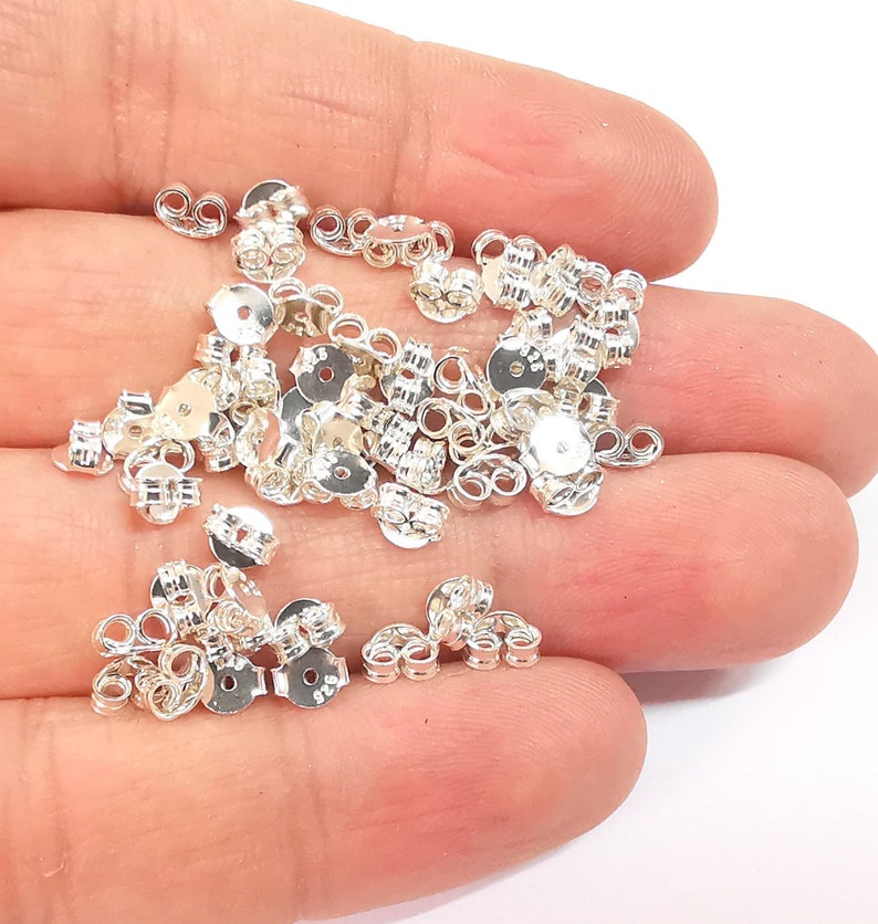 May include: A pile of silver earring backs. The backs are made of metal and have a circular shape with a small hole in the center. They are designed to be used with stud earrings.