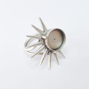 May include: A silver-colored ring with a sunburst design. The ring features a circular setting in the center, surrounded by pointed rays. The band is a double-loop design. The ring is set against a white background.