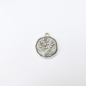 May include: A silver-colored round pendant featuring a detailed relief of lily of the valley flowers and leaves. The pendant has a small loop at the top for attaching to a chain or cord. The design is set against a textured background.