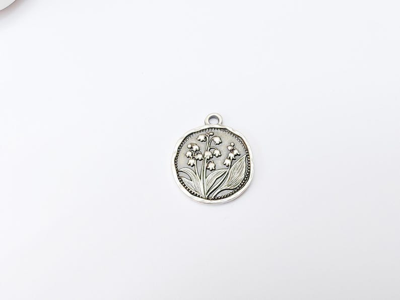 May include: A silver-colored round pendant featuring a detailed relief of lily of the valley flowers and leaves. The pendant has a small loop at the top for attaching to a chain or cord. The design is set against a textured background.