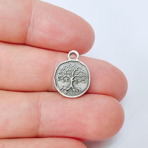 May include: A small, round, silver-colored charm featuring a detailed tree design. The tree has intricate branches and roots, set within a circular frame with a small loop at the top for attaching to jewelry or other crafts.