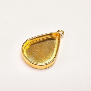 Pode incluir: Um suporte de pingente em forma de gota de cor dourada, com uma moldura elevada e um pequeno laço para prender uma corrente ou cordão. O pingente tem um acabamento liso e polido e foi projetado para conter um cabochão ou outro elemento decorativo.