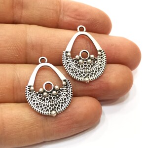 May include: Two silver-toned metal earring charms with a delicate, intricate design. Each charm features a teardrop shape with a circular opening at the top and a decorative, scalloped bottom.