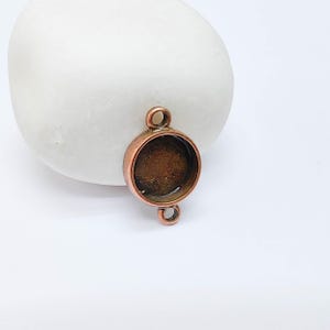 May include: A copper-colored round bezel connector with two loops and a circular setting. The jewelry component is designed for crafting and jewelry making. The bezel is empty, ready to hold a stone or other embellishment.