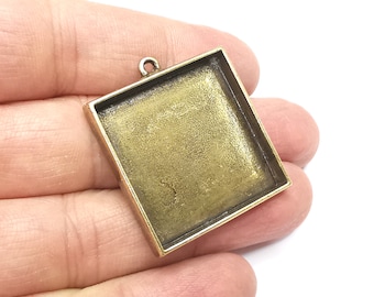 May include: Square antique bronze pendant tray with a loop for attaching to a chain. The pendant has a recessed square area for adding a design or image. The pendant is held in a hand against a white background.