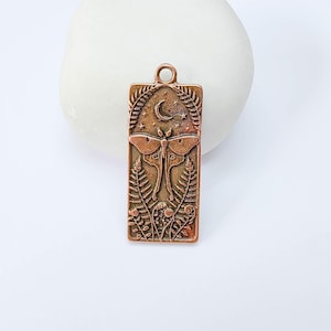Puede incluir: Un colgante rectangular de metal en tono cobre con un diseño en relieve de una polilla sobre frondas de helecho. La parte superior muestra una luna creciente y estrellas pequeñas. Incluye una anilla circular en la parte superior para colgar.