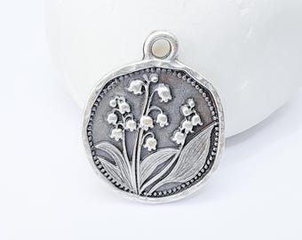 Puede incluir: Colgante redondo plateado con un relieve detallado de flores y hojas de lirio de los valles. El colgante tiene un borde texturizado y un pequeño lazo en la parte superior para una cadena. El diseño está centrado sobre un fondo blanco.
