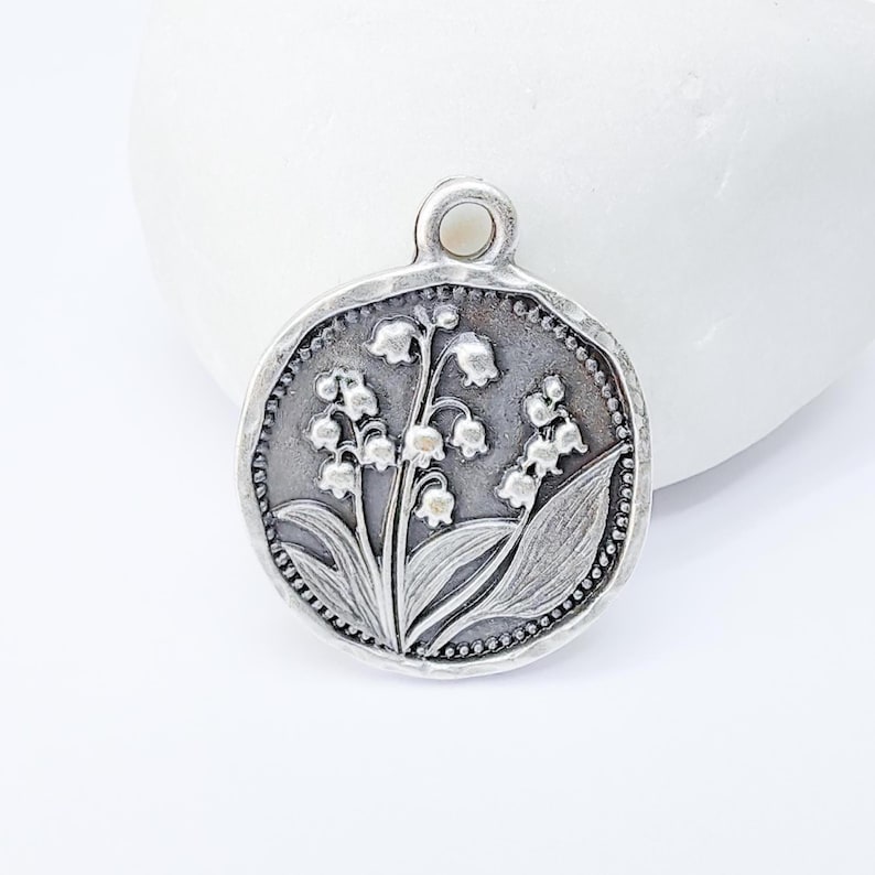 May include: A round, silver-toned pendant featuring a detailed relief of lily of the valley flowers and leaves. The pendant has a textured border with a small loop at the top for a chain. The design is centered on a white background.