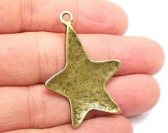 Peut inclure: Un charm en forme d'étoile de couleur bronze avec une surface texturée. Le charm a une boucle en haut pour l'attacher à un collier ou un bracelet. L'étoile a cinq pointes, dont une allongée. Le charm est tenu dans la main d'une personne.