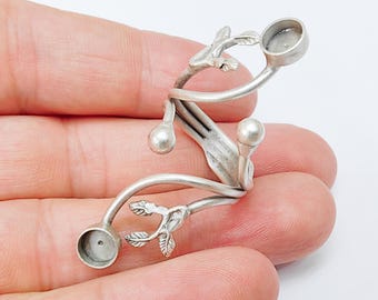 Puede incluir: Un pendiente de oreja plateado con un diseño botánico. El pendiente presenta líneas curvas, pequeñas hojas y dos engastes circulares para sujetar pendientes. El diseño se sostiene en una mano sobre un fondo blanco.