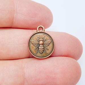 May include: A round, copper-colored charm with a raised bee design. The bee is centered on the charm, with detailed wings and body. The charm has a small loop at the top for attaching to a chain or other jewelry. The charm is surrounded by a dotted border.