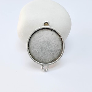 May include: A round, silver-toned pendant base with a bezel setting. The pendant has two small loops for attaching to a chain or other jewelry components. The setting is empty, ready for a cabochon or other insert.