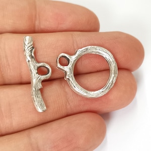May include: Silver tone metal toggle clasp with a twig design. The clasp has two holes for attaching to a necklace or bracelet.