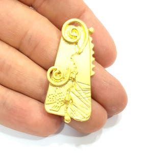 May include: A gold-toned pendant with a geometric design and intricate swirls. The pendant has a textured surface and a slightly irregular shape.