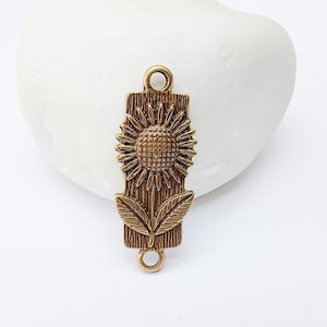 Op de afbeelding: Een bronzen metalen bedel met een zonnebloemontwerp. De bedel heeft een rechthoekige vorm met een zonnebloem in het midden en bladeren aan de onderkant. Het heeft lussen aan de boven- en onderkant om aan sieraden te bevestigen.