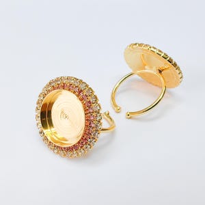 May include: Two gold-coloured adjustable rings with a round, flat top. The top is surrounded by a row of small, clear and pink rhinestones. The ring base is a simple, open design. The rings are displayed on a white background.