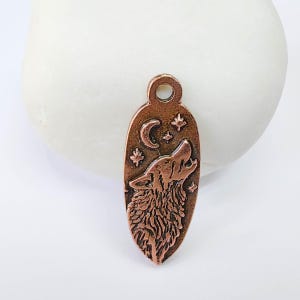 May include: A copper-colored pendant with a howling wolf design. The oval-shaped pendant features a wolf howling at the moon and stars. The pendant has a small hole at the top for attaching to a chain or cord.