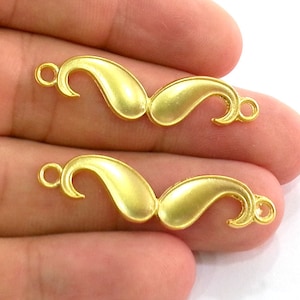 May include: Two gold-colored metal paisley-shaped charms with a loop on each end. The charms are on a hand.