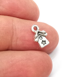 May include: Silver charm in the shape of a small house with a star on the front. The charm is being held by a finger.