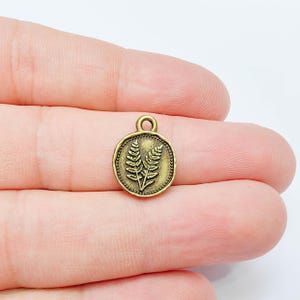 May include: A small, round, antique brass-toned metal charm featuring an embossed fern leaf design on the front. The circular charm has a beaded border and a small loop at the top for attachment, presented against a plain white background.