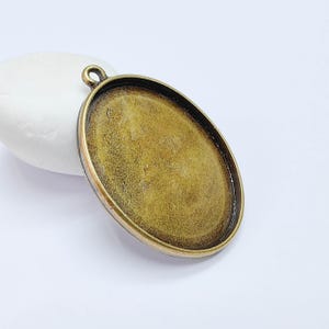 Könnte beinhalten: Ein ovaler Anhänger mit Bronze-Finish. Er hat oben eine Schlaufe zum Befestigen an einer Kette oder einem Band. Der Innenraum ist eine vertiefte Fläche zum Hinzufügen eines Designs oder Bildes. Der Anhänger liegt auf einer weißen Oberfläche.