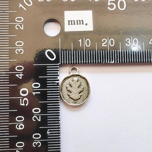 以下が含まれることがあります： 円形のフレーム内に、浮き彫りのオークの葉のデザインが施された小さな銀色のペンダント。ペンダントの上部には、チェーンなどに取り付けるためのループが付いています。ミリメートル単位の目盛りが付いた定規の隣にあります。直径は約1cmです。