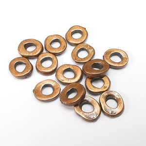 Pode incluir: Uma coleção de contas em forma de anel de cor bronze. Cada conta tem um buraco no centro e uma superfície ligeiramente irregular e texturizada. As contas estão espalhadas sobre um fundo branco, mostrando seu brilho metálico e formas únicas.
