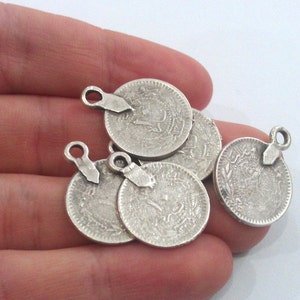 May include: Five silver-toned coin charms with a loop for hanging. Each charm features an intricate design and a raised edge.